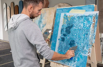 Un partecipante applica con la spatola una finitura azzurra attraverso uno stencil a piastrelle